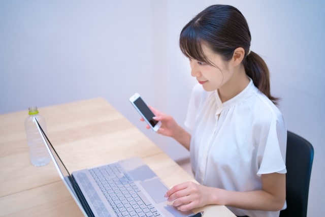 パソコンを見ながらスマホを操作する女性の画像